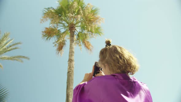 a Young European Girl in a Vintage Dressing Gown is Taking Pictures of a Tall Palm Tree Crashing