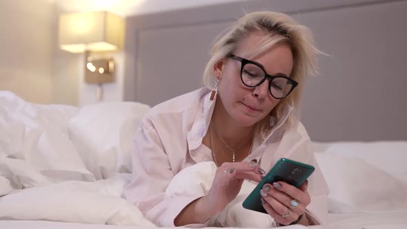 Closeup of a Blonde Woman with Glasses Is in the Bedroom Lying on Her Stomach on a Bed with White