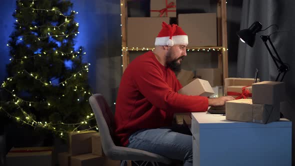 Happy Idus Bearded Santa Claus in a Suit Sits in a Home Office at a Table Among Boxes Makes