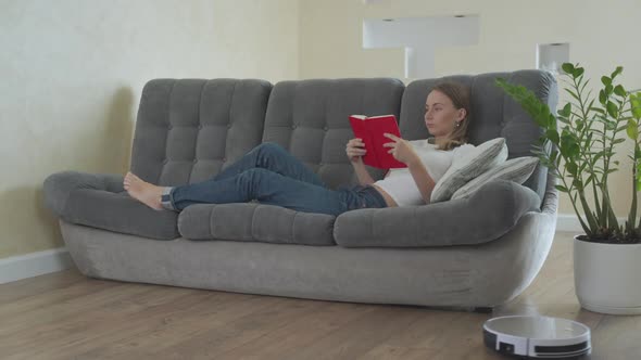 Woman Reading Book While Robotic Vacuum Cleaner Cleaning House