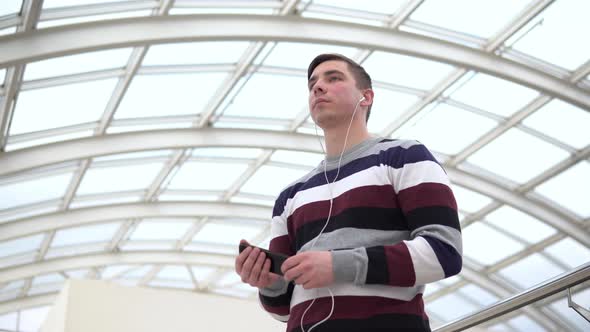A Young Man Is Standing in Headphones. A Man Listens To Music on Headphones Against a Pan-glass Roof
