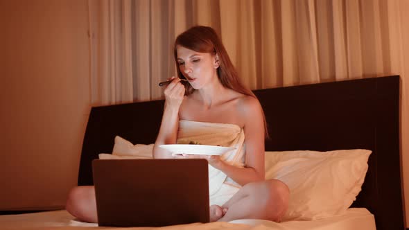 Young Woman Sitting on Bed with Plate of Tasty Dish and Watching Movie on Modern