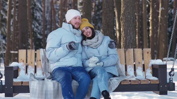 Young Couple Rests in a Winter Forest Man and a Young Woman Sit on a Swing and Relaxing in a Winter
