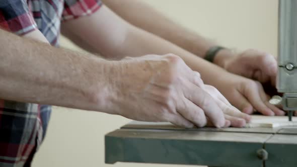 Tight panning shot of grandfather's hands helping and guiding grandson cut wood with an band saw.