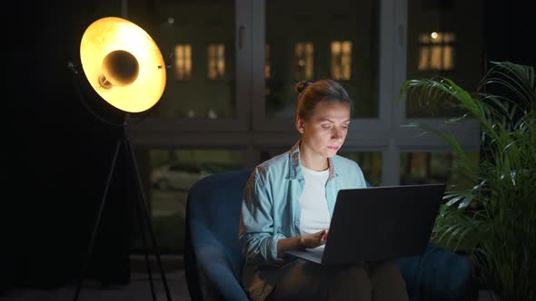 Woman is Sitting in the Armchair and Working on a Laptop at Night or Texting Someone