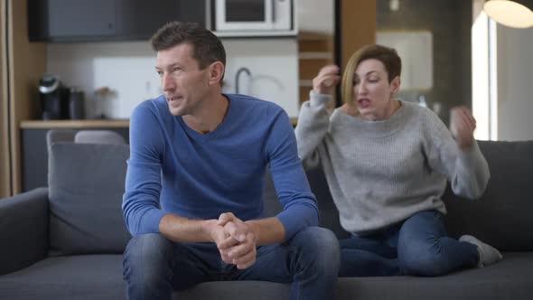 Portrait of Sad Stressed Caucasian Man Sitting on Couch with Woman Yelling Gesturing Arguing with