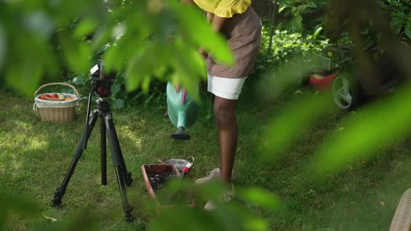 Unrecognizable African American Blogger Watering Seedlings in Pot Filming Video Blog in Garden