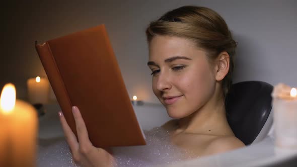 Young Woman Reading Book in Bath With Foam and Candles, Evening Relaxation