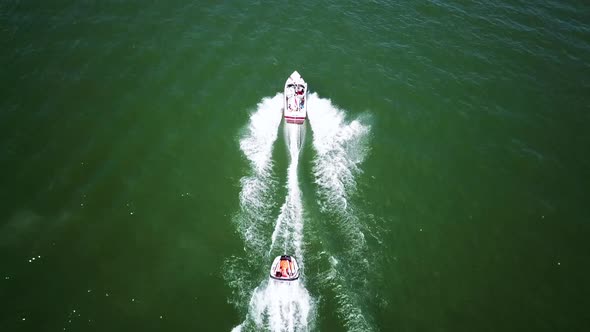 Drone Shot of a boat on a green lake pulling behind it a tube with people on it, creating a large wa