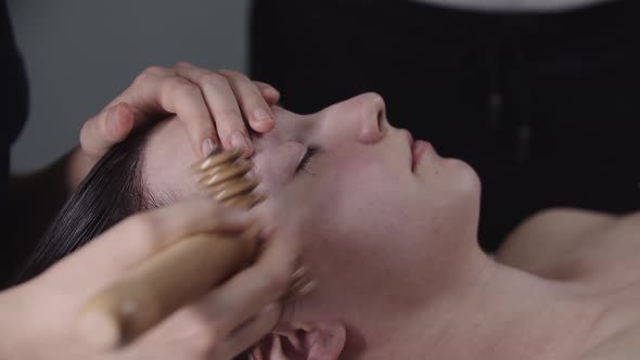 Woman Massagist Using a Wooden Ribbed Roller Tool on the Cheek of Her Female Client