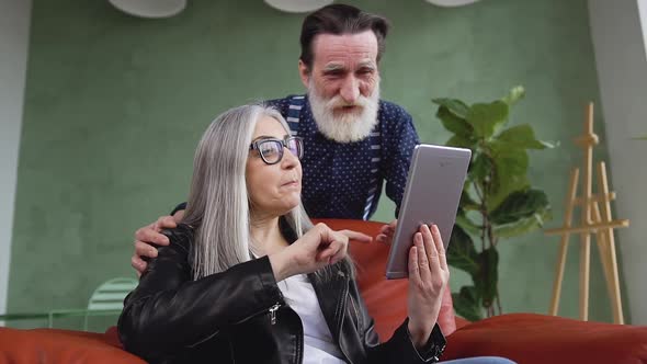 Modern Mature Woman and Her Bearded Husband Using Tablet PC and Talking