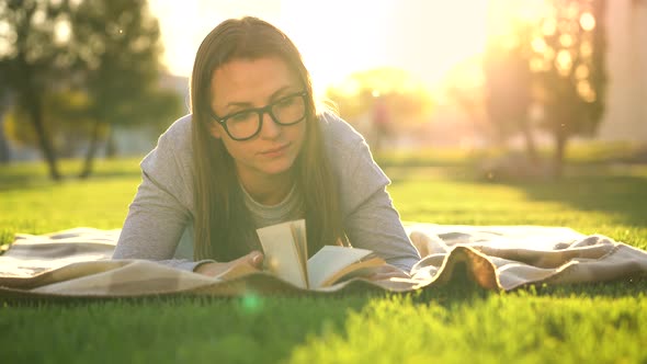 Girl in Glasses Reading Book Lying Down on a Blanket in the Park at Sunset