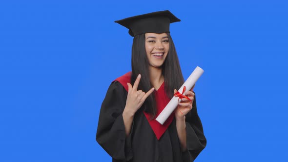 Portrait of Female Student in Cap and Gown Graduation Costume Holding Diploma and Making Rock