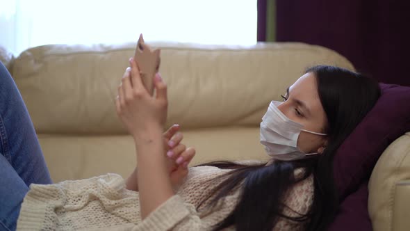 Woman in a Medical Mask Uses a Smartphone at Home During Quarantine.