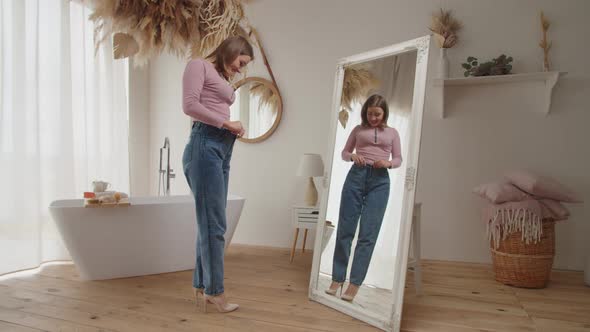 Cheerful Slim Adult Woman in Loose Jeans Checking in Mirror Her Weight Loss Success at Home