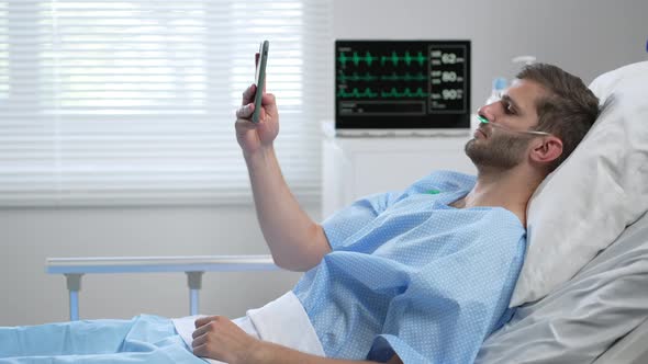 A Young Male Clinic Patient is Lying on a Hospital Bed and Calls Via Video Link Using a Mobile Phone