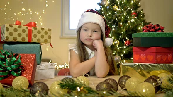 Little Girl in a Santa Hat Sits Sadly with Her Head on Her Hand