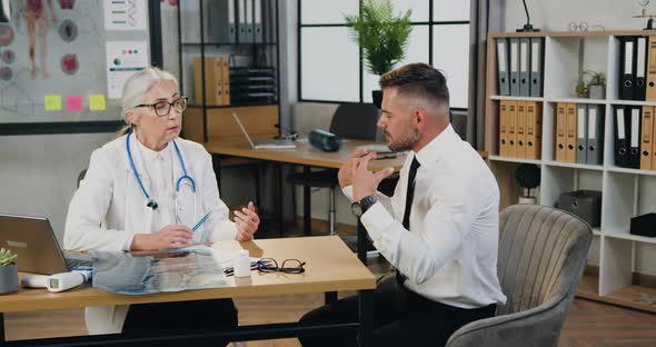 Man Visiting Medical Office and Telling Caring Professional Mature Grey-Haired Female Doctor