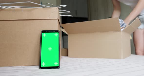 Phone green screen mockup near box, woman packing pre owned clothes.