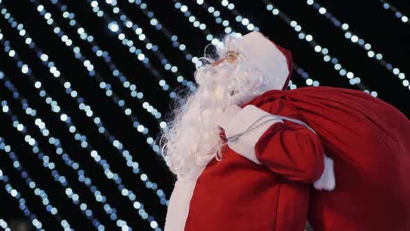 Santa Claus Looks Up at the City Christmas Tree and Bright Flashing Lights of Garlands