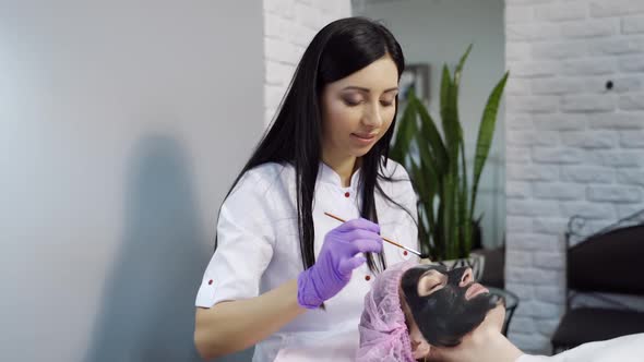 A beautiful cosmetologist applies a mask to clean the pores on the face of the client