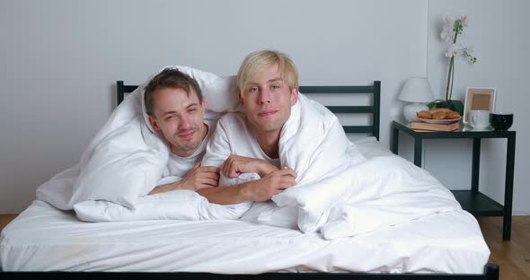 Young Gay Couple Lying on Bed Together in Bedroom Hugging and Kissing