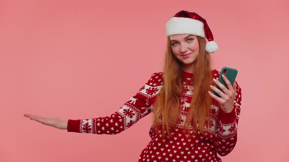 Girl in Christmas Santa Sweater with Mobile Phone Showing Pointing Empty Place Advertising Area