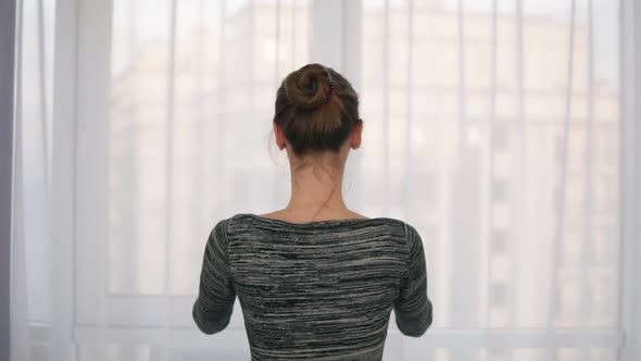 Back View of Young Woman Coming Up to the Window Unveiling Curtains and Looking Out of Window