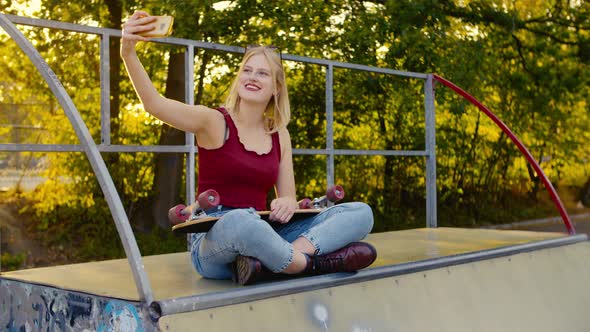 Happy fit skater Girl is taking a selfie sitting on a halfpipe in the sun,With the sun behind her b