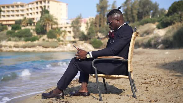 Confident Stylish African American Man Sitting on Wicker Chair Surfing Internet on Tablet Admiring