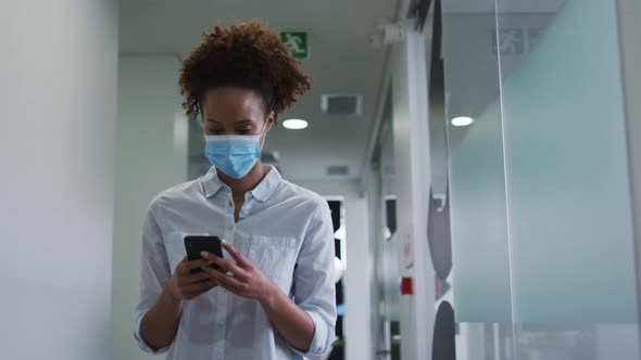 Mixed race businesswoman in face mask using smartphone walking in corridor in office