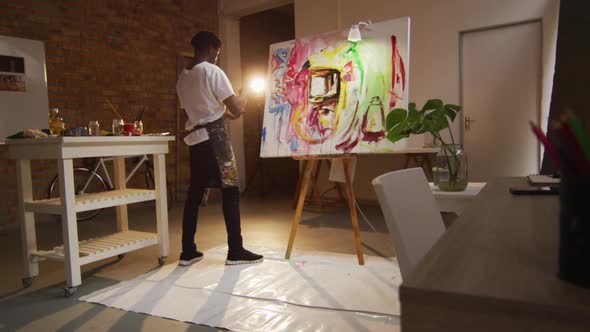 African american male artist wearing apron painting with palette knife on canvas at art studio