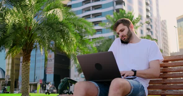 A Man in a White T-shirt with a Laptop Talking on the Phone in the Summer on the Bench