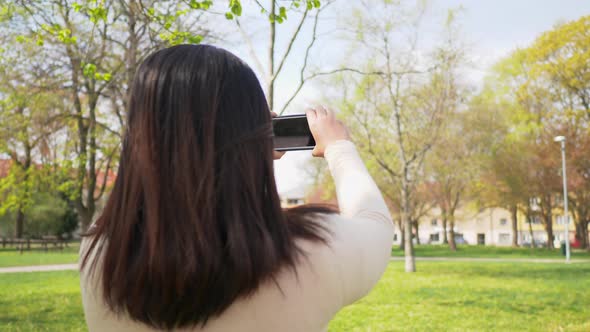 Back view of Asian woman standing at a park and taking a photo of park view on the weekend