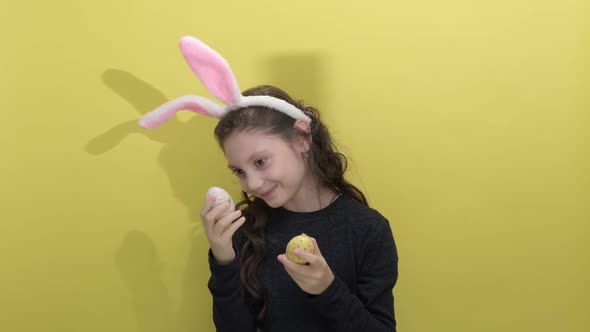 Little Girl in Easter Bunny Ears Holding Two Painted Easter Eggs on a Yellow
