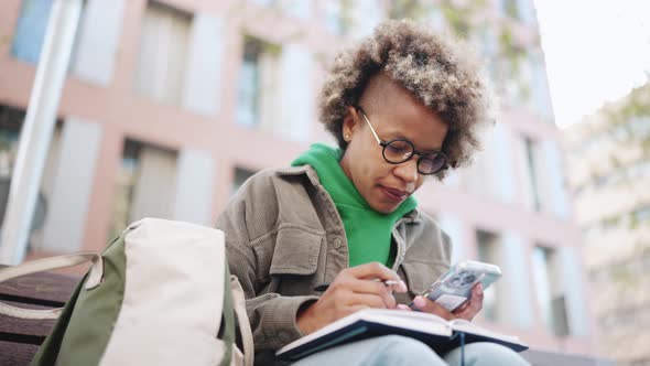 Concentrated African woman wearing green hoodie writing notes in notepad from smartphone