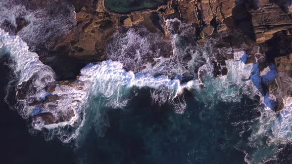 Top bird's eye view of ocean blue waves crashing against rocky Australian coastline and rockpool dro