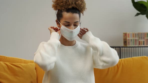 A Black Woman Puts a Mask on Her Face While Sitting on a Yellow Sofa at Home During a Virus Pandemic