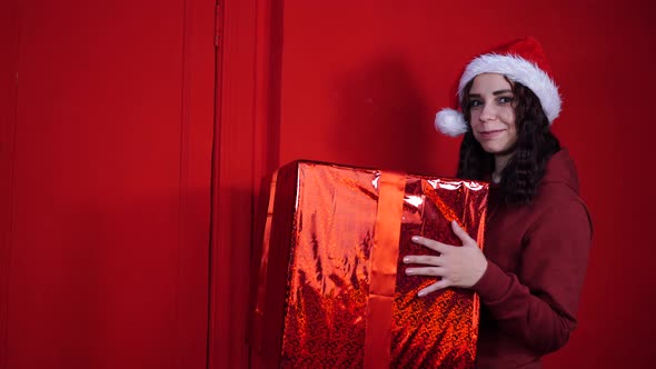 Young Woman in Santa Claus Hat Holds Big Gift Standing Near Red Wall