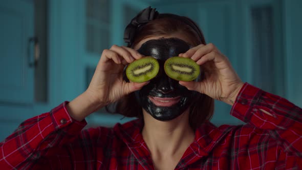 30s woman with black cosmetic mask on her face holding kiwi fruit halves near eyes