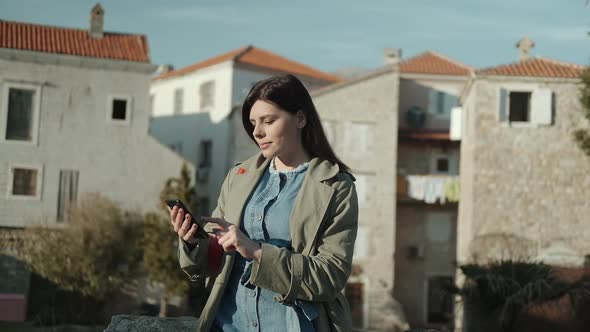 Pretty young woman with brunette hair in green coat  texting on her phone  in old town in Montenegro