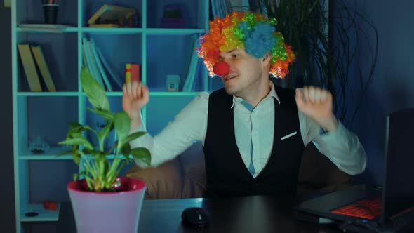 A Young Man in the Image of a Clown Dances in the Workplace Sitting at a Computer in the Office