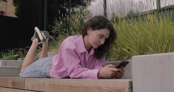 Smiling white woman in city, lying on deck wearing headphones and using smartphone in park.