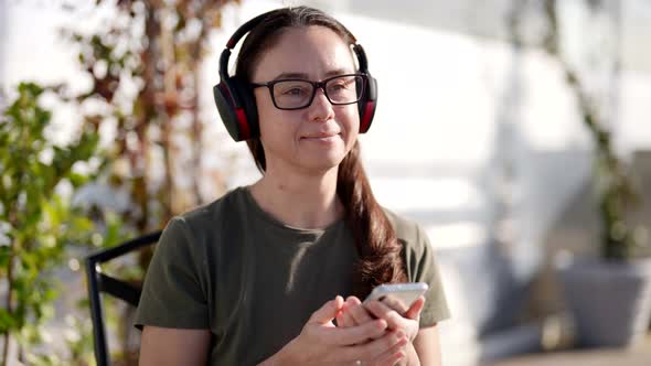 An Attractive Adult Woman in Stylish Glasses and Wireless Headphones Listens to a Radio Program in a