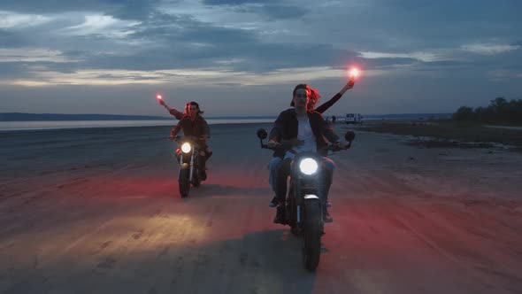 Two Young Couples Riding on Vintage Motorcycles with Red Burning Signal Flares After Sunset on Beach