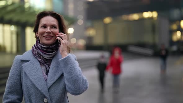 a Woman in a Light Coat and Scarf and with Bright Nails is Talking on the Phone and Laughing at Dusk