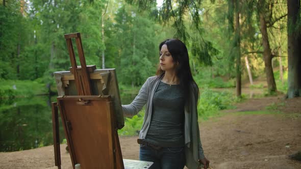 Beautiful Girl Artist in a Blouse and Jeans Paints a Landscape in a Park Near a Pond