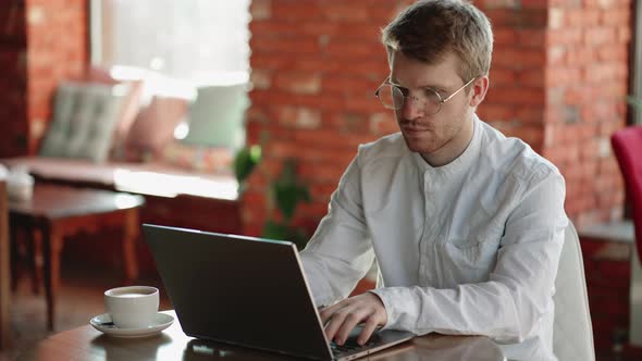 Concentrated Young Hipster Freelancer Man with Beard in Glasses Sitting at the Table and Looking at