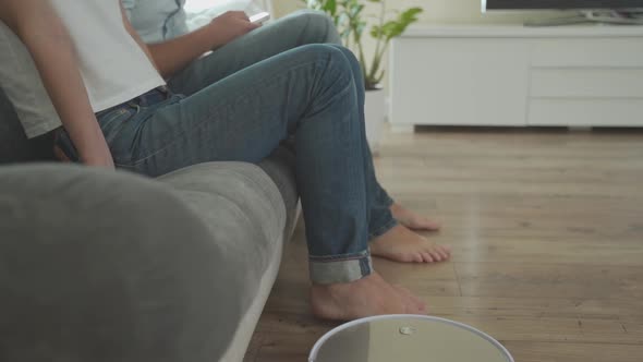 Portrait of Lovely Couple Relaxing on Sofa and Lifting Up Their Feet While Automatic Vacuum Cleaner