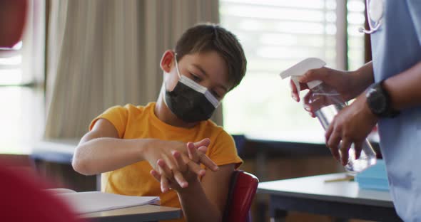 Portrait of mixed race schoolboy wearing face masks, disinfecting hands in classroom